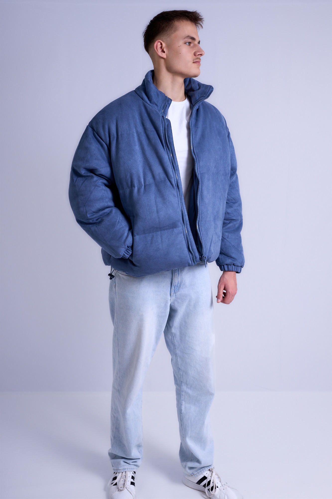 Junger Mann in blauer Daunenjacke und hellen Jeans, steht lässig mit neutralem Ausdruck.