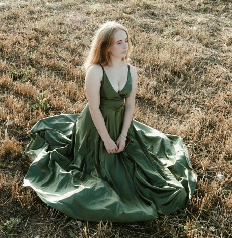 Frau in grünem Kleid sitzt auf einer trockenen Wiese, umgeben von hohem Gras.
