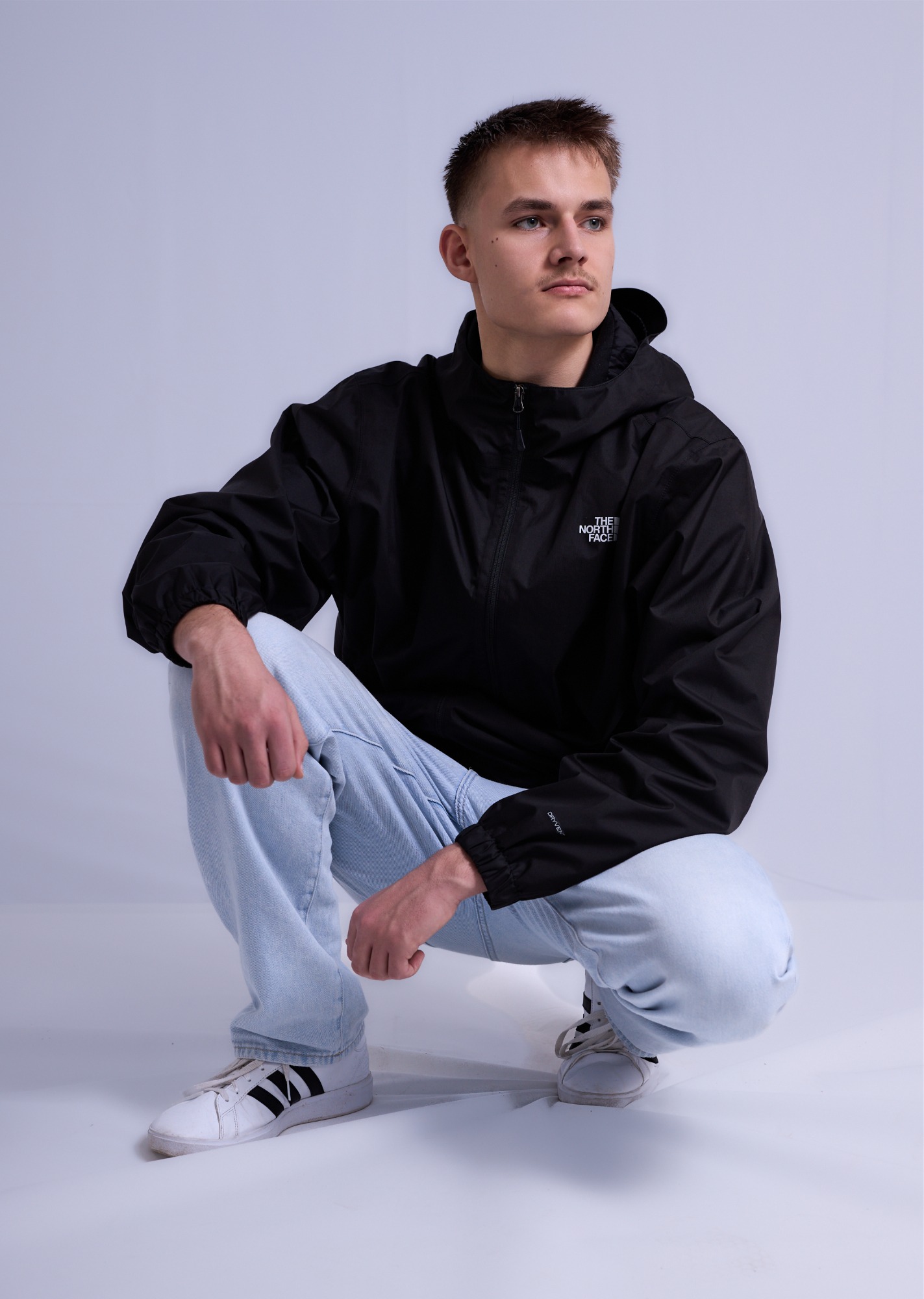 Junger Mann in schwarzer Jacke und hellblauen Jeans, hockend auf einem hellen Hintergrund.