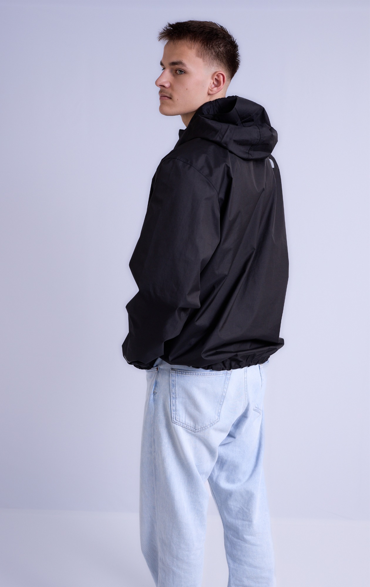 Junger Mann in schwarzer Jacke und hellen Jeans, von hinten mit neutralem Hintergrund.