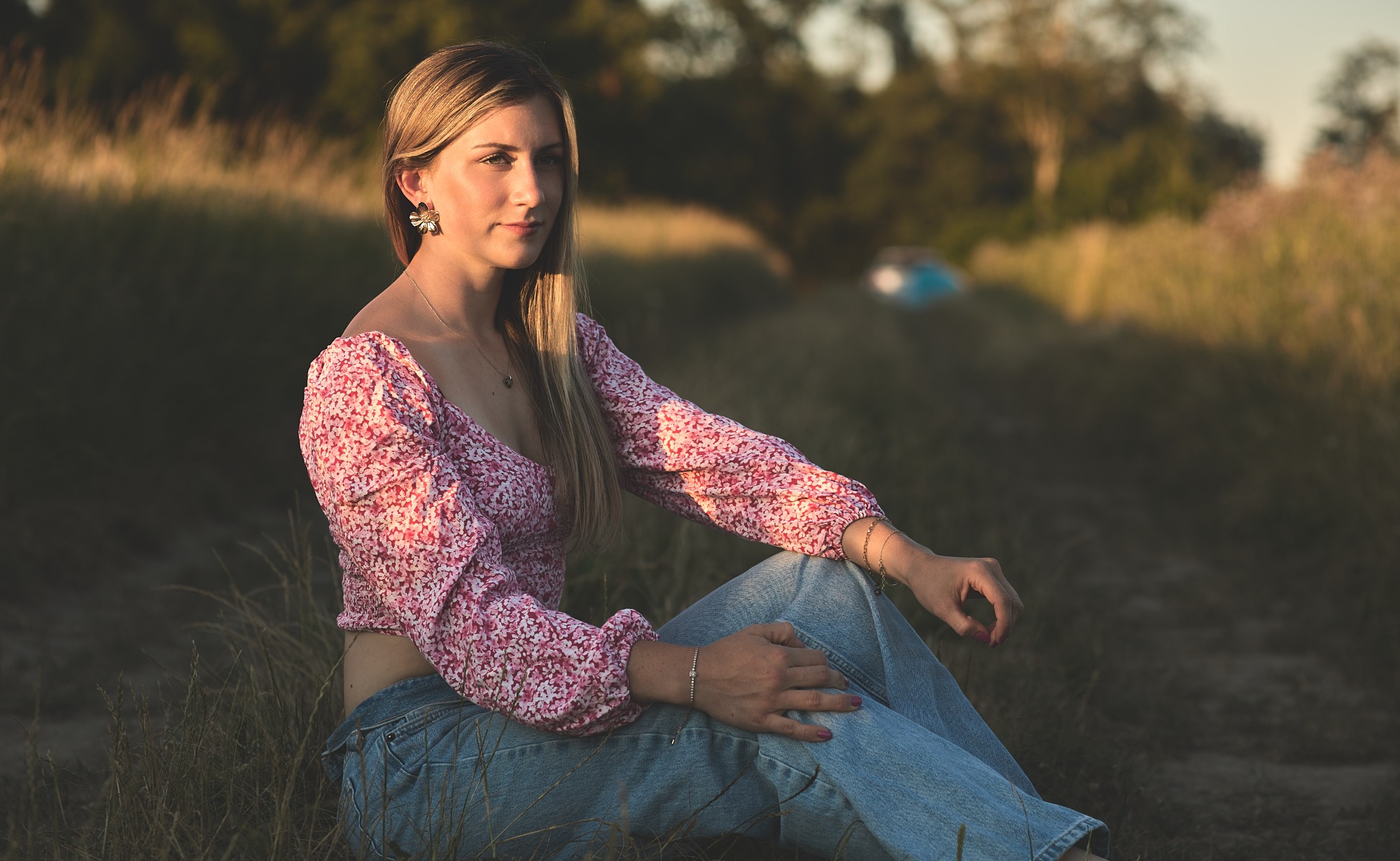 Junge Frau im Freien, sitzend auf dem Boden, trägt ein rosa Oberteil und blaue Jeans.