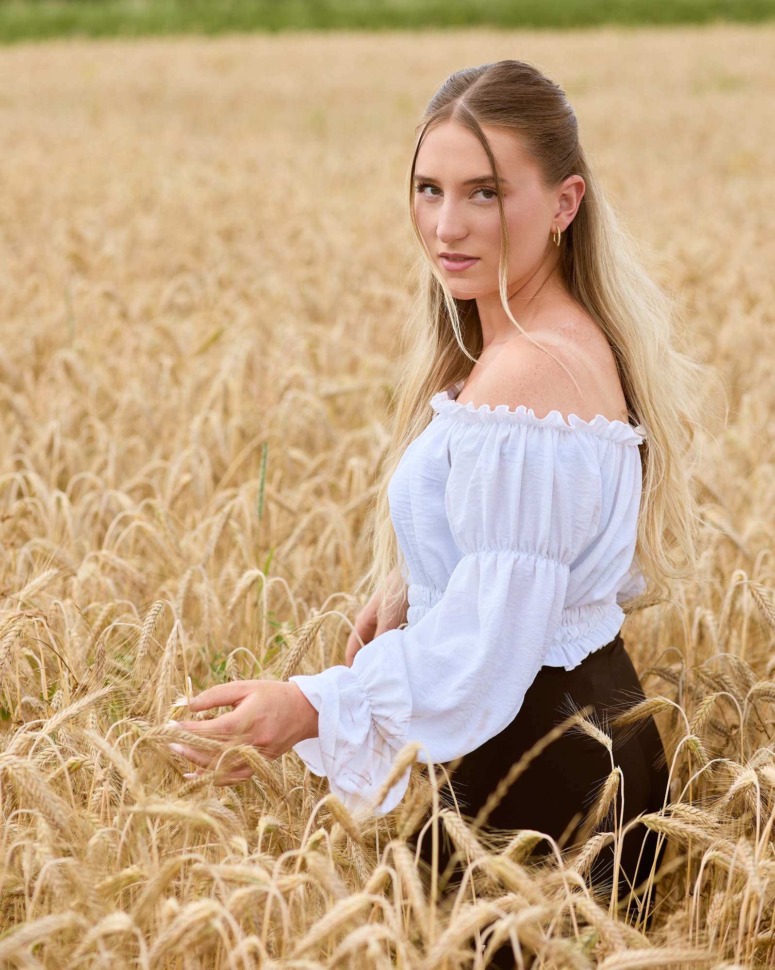 Junge Frau in einem Weizenfeld, trägt ein off-shoulder Oberteil und blickt direkt zur Seite.