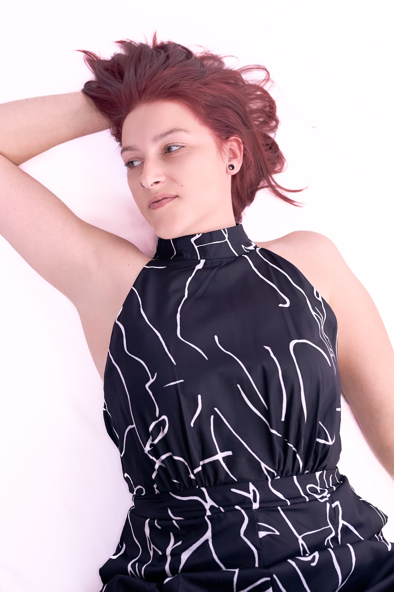 Junge Frau mit rotem Haar, in elegantem schwarzen Kleid, entspannt auf hellem Hintergrund.